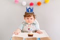 Cute adorable Caucasian baby boy in blue crown celebrating first birthday at home. Child kid toddler sitting in high chair looking Royalty Free Stock Photo