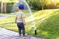 Cute adorable caucasian blond barefeet toddler boy in cap walking at home backyard near sprinkler automatic watering