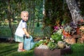 Cute adorable caucasian blond little toddler boy in watering flowerbed flower pot with green plastic can outdoors. Fun baby boy