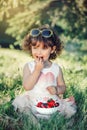 Caucasian toddler baby girl sitting on grass and eating berries fruits. Funny child with sunglasses in park having healthy snack