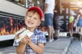 Cute adorable little happy smiling boy kid enjoy eating hot dog sausage in bread near street cafe stall outdoors. Child Royalty Free Stock Photo