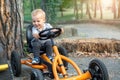 Cute adorable smiling caucasian toddler boy sitting at pedal toy car and hold, steering wheel by hands driving it outdoors in city