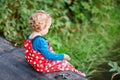 Cute adorable toddler girl sitting on wooden bridge and throwing small stones into a creek. Funny baby having fun with Royalty Free Stock Photo