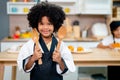 Cute African American girl hold wooden spoon and fork and stand in front of table in kitchen during cooking class Royalty Free Stock Photo