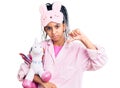 Cute african american girl wearing sleep mask and pajama holding pink teddy unicorn with angry face, negative sign showing dislike