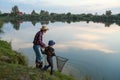 Cute 10-aged boy with his experienced grey-bearded 70-aged grandpa catching fish on the lake with landing net at sunset Royalty Free Stock Photo