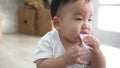 Cute Asian baby biting piece of snack while sitting on floor in cozy room at home