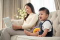 A cute Asian baby boy is playing with toys on a sofa while his mom is working on her laptop Royalty Free Stock Photo