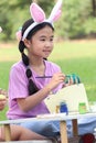 Cute Asian girl with bunny ears painting eggs with paintbrush while sitting on green grass meadow in nature garden. Kid Royalty Free Stock Photo