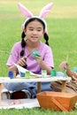 Cute Asian girl with bunny ears painting eggs with paintbrush while sitting on green grass meadow in nature garden. Kid Royalty Free Stock Photo
