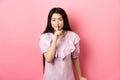 Cute asian girl hiding secret, hushing with finger pressed to lips and smiling, asking to keep quiet, standing in dress Royalty Free Stock Photo