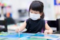 Cute Asian girl wearing cloth face mask while taking art classes in class. Little child painted the water on the canvas.