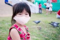 Cute Asian girl wearing mask sitting on grass. Behind the little child was flock of pigeons on green lawn. Royalty Free Stock Photo