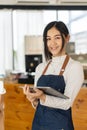 Cute Asian woman holding a tablet in an apron standing in front of a coffee shop, customer service concept. Royalty Free Stock Photo