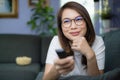 Cute Asian woman laying on sofa with relax and cozy gesture holding remote control aiming to tv, watching television and smile