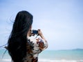Cute asian woman wearing retro dress standing at seaside holding camera in hand and take a photo Royalty Free Stock Photo