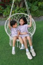 Cute Asian young sister and little brother looking at each other and sitting together on the white cradle in the garden. Boy and Royalty Free Stock Photo