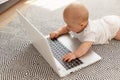 Cute baby being interested in modern technology, lying on floor and using laptop computer why mother are gone, infant girl on gray Royalty Free Stock Photo