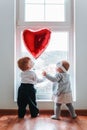 Cute baby boy and baby girl looking at the red heart balloon. Window at the background. The concept of Valentine`s day Royalty Free Stock Photo