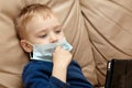 Cute baby boy with blond hair and blue eyes in medical sanitary mask sitting on beige leather sofa with pad digital tablet. Wearin Royalty Free Stock Photo