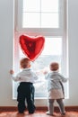 Cute baby boy and baby girl looking at the window. Boy holding a red heart balloon. Rear view. The concept of Valentine`s day Royalty Free Stock Photo