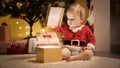 Cute baby boy sitting under Christmas tree and opening lid of golden gift box Royalty Free Stock Photo