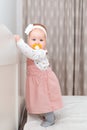 A cute baby girl in a dress, with a pacifier in her mouth and with a headband on her head, stands near the crib. The concept of a Royalty Free Stock Photo