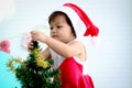 Cute baby girl kid in Santa Claus red costume trying to decorate Christmas tree in white living room, beautiful little child Royalty Free Stock Photo