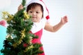 Cute baby girl kid in Santa Claus red costume trying to decorate Christmas tree in white living room, beautiful little child Royalty Free Stock Photo