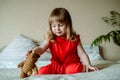 Cute baby girl in a red dress plays on the bed in a bright bedroom. Happy childhood. Teddy bear. joy and laughter of a child Royalty Free Stock Photo