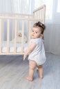 Cute baby girl stands at the side of the crib holding on to it with her hands in a white bodysuit, baby learns to walk Royalty Free Stock Photo