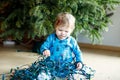 Cute baby girl taking down holiday decorations from Christmas tree. child holding light garland. Royalty Free Stock Photo