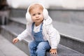 Cute baby girl 1-2 year old wear fluffy white headphones, denim suit pants and knitted sweater posing on city street outdoors. Royalty Free Stock Photo