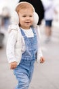 Cute baby girl 1-2 year old wear fluffy white headphones, denim suit pants and knitted sweater posing on city street outdoors. Royalty Free Stock Photo