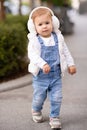 Cute baby girl 1-2 year old wear fluffy white headphones, denim suit pants and knitted sweater posing on city street outdoors. Royalty Free Stock Photo