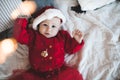 Cute baby girl 1 year old wear red santa claus hat and knit dress lying in bed close up. Smiling little child celebrating Royalty Free Stock Photo