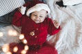 Cute baby girl 1 year old wear red santa claus hat and knit dress lying in bed close up. Smiling little child celebrating Royalty Free Stock Photo