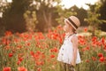 Cute baby girl 4-5 year old wear white stylish dress and straw hat standing in poppy flower in field outdoors. Royalty Free Stock Photo