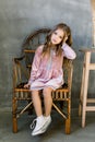 Cute baby girl 5-6 year old wearing stylish pink dress sitting on the wooden chair overgrey background. Looking at Royalty Free Stock Photo