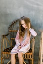 Cute baby girl 5-6 year old wearing stylish pink dress sitting on the wooden chair overgrey background. Looking at Royalty Free Stock Photo