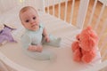 Cute baby 9 months old sits in an oval white crib with a toy bear.