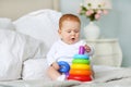 Cute baby playing with colorful rainbow toy pyramid sitting on bed in white sunny bedroom. Toys for little kids. Royalty Free Stock Photo