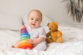 Cute baby playing with colorful rainbow toy pyramid and teddy bear sitting on bed in white sunny bedroom copy space Royalty Free Stock Photo