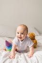 Cute baby playing with colorful rainbow toy pyramid and teddy bear sitting on bed in white sunny bedroom. Toys for Royalty Free Stock Photo