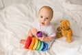 Cute baby playing with colorful rainbow toy pyramid and teddy bear sitting on bed in white sunny bedroom. Toys for Royalty Free Stock Photo