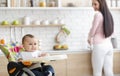 Cute baby playing on high chair in kitchen, mother washing dishes