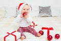 Cute baby with red Santa Claus hat and gift box taste red christmas ball on white bed. Christmas congratulations concept with copy Royalty Free Stock Photo