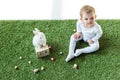 Baby sitting on green grass near wooden calendar with 28 April date, decorative rabbit and colorful quail eggs isolated Royalty Free Stock Photo