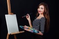 Cute beautiful girl artist painting a picture on canvas an easel. Space for text. Studio black background. Royalty Free Stock Photo