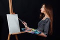 Cute beautiful girl artist painting a picture on canvas an easel. Space for text. Studio black background. Royalty Free Stock Photo